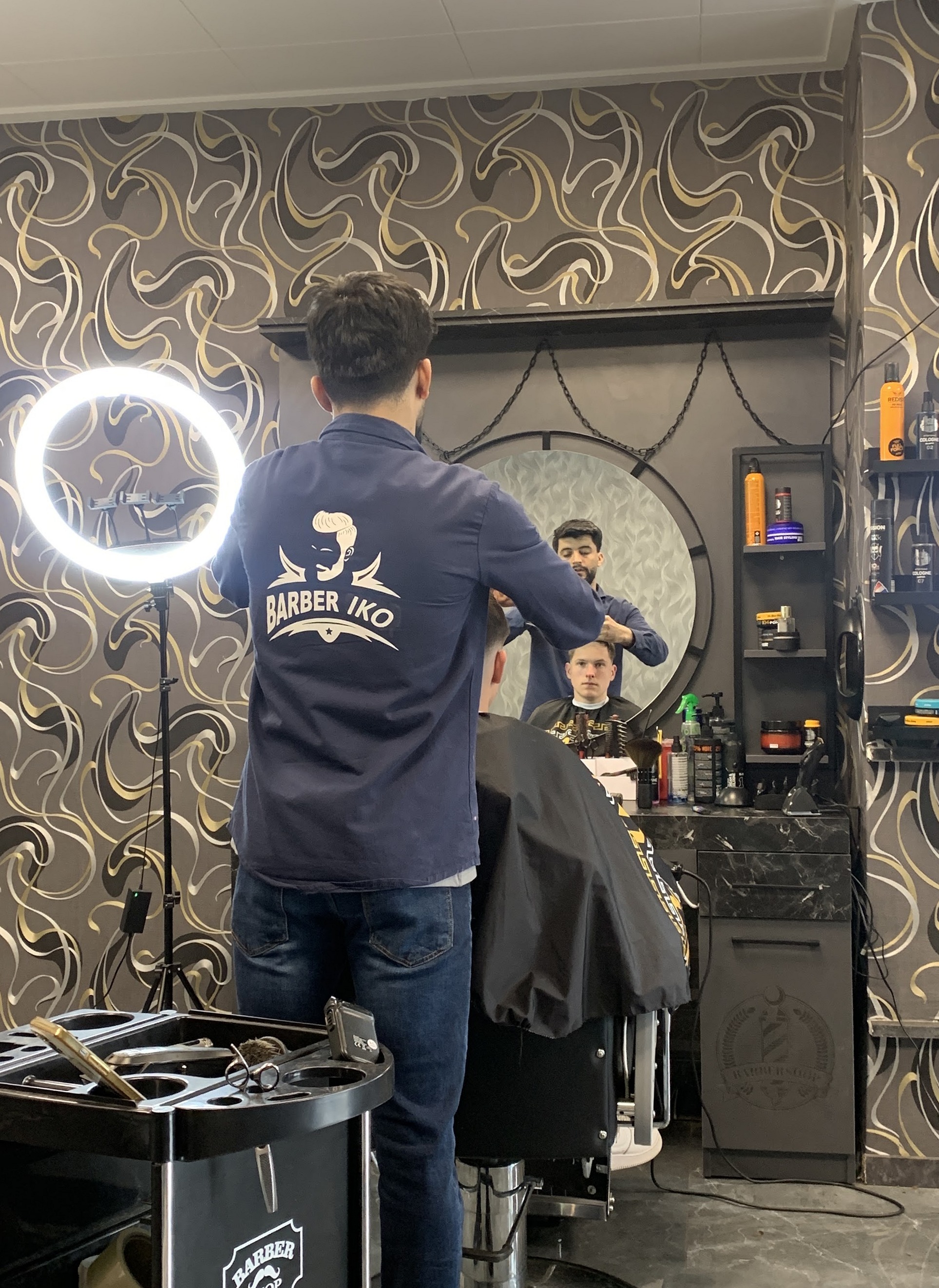 Een barber in het navy Barber IKO shirt aan het werk voor de ronde spiegel, met de ringlamp ernaast